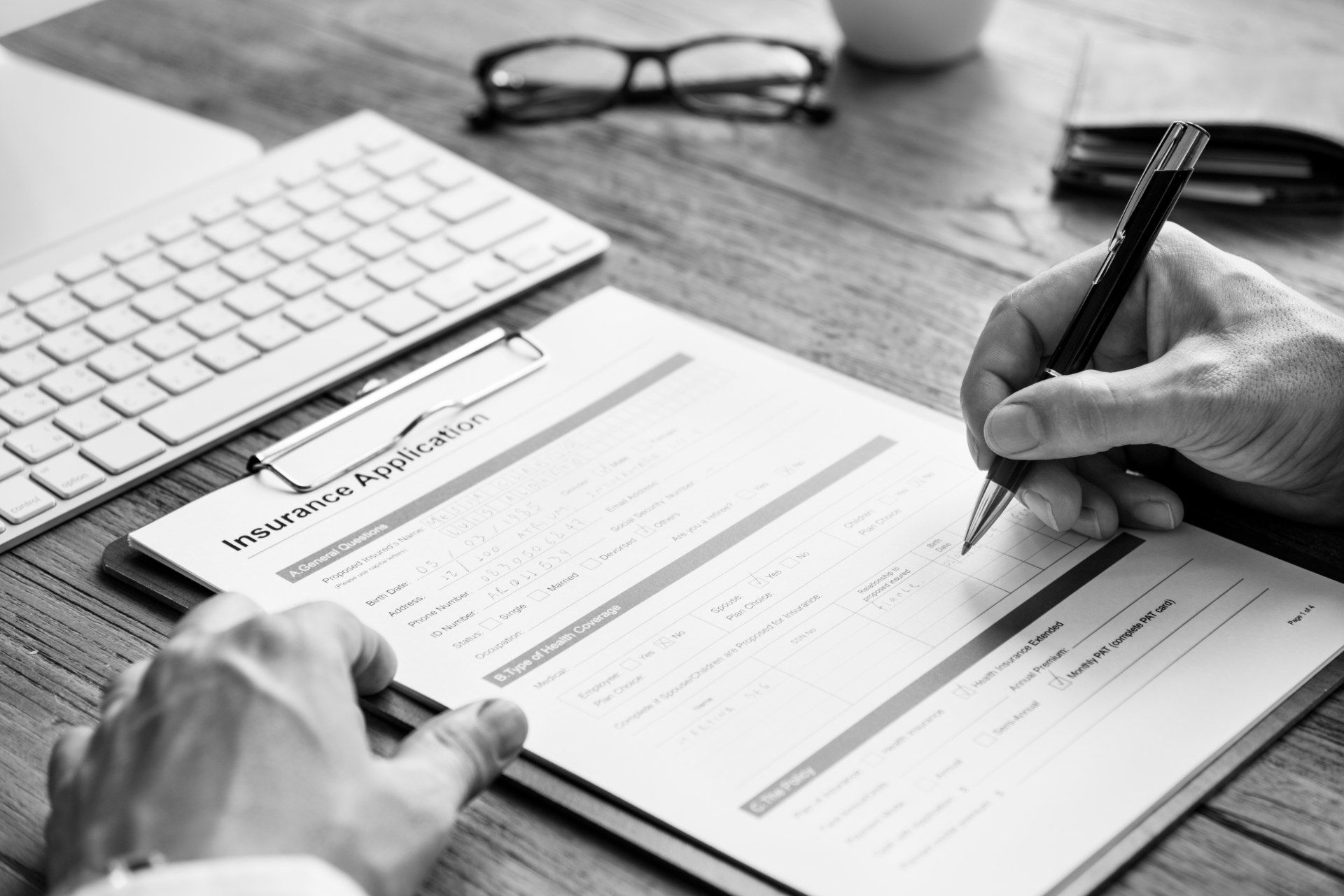 Person fills out insurance form on clipboard next to keyboard and reading glasses - insurance premiums
