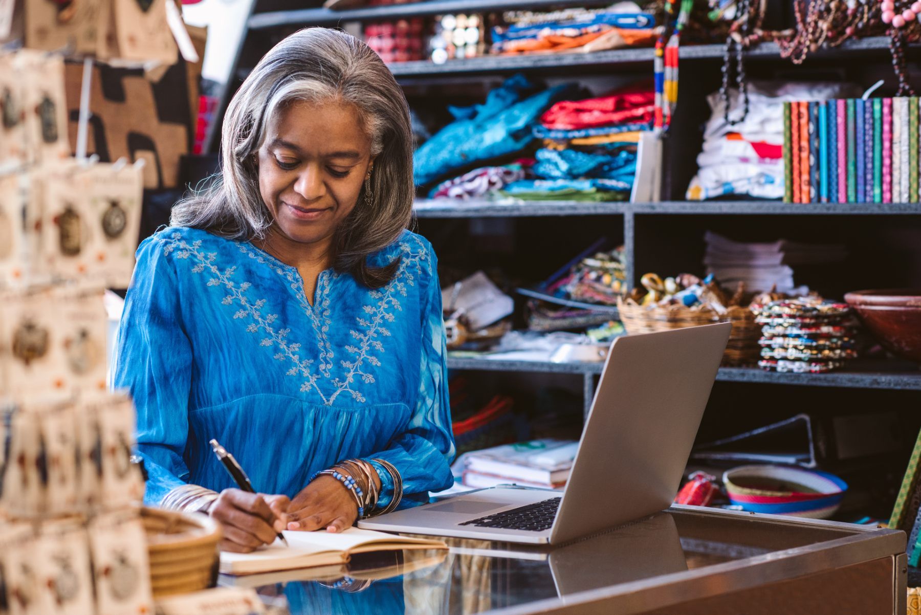 A woman writes in a notebook near her laptop on the counter at a fabric store - self-employment grant