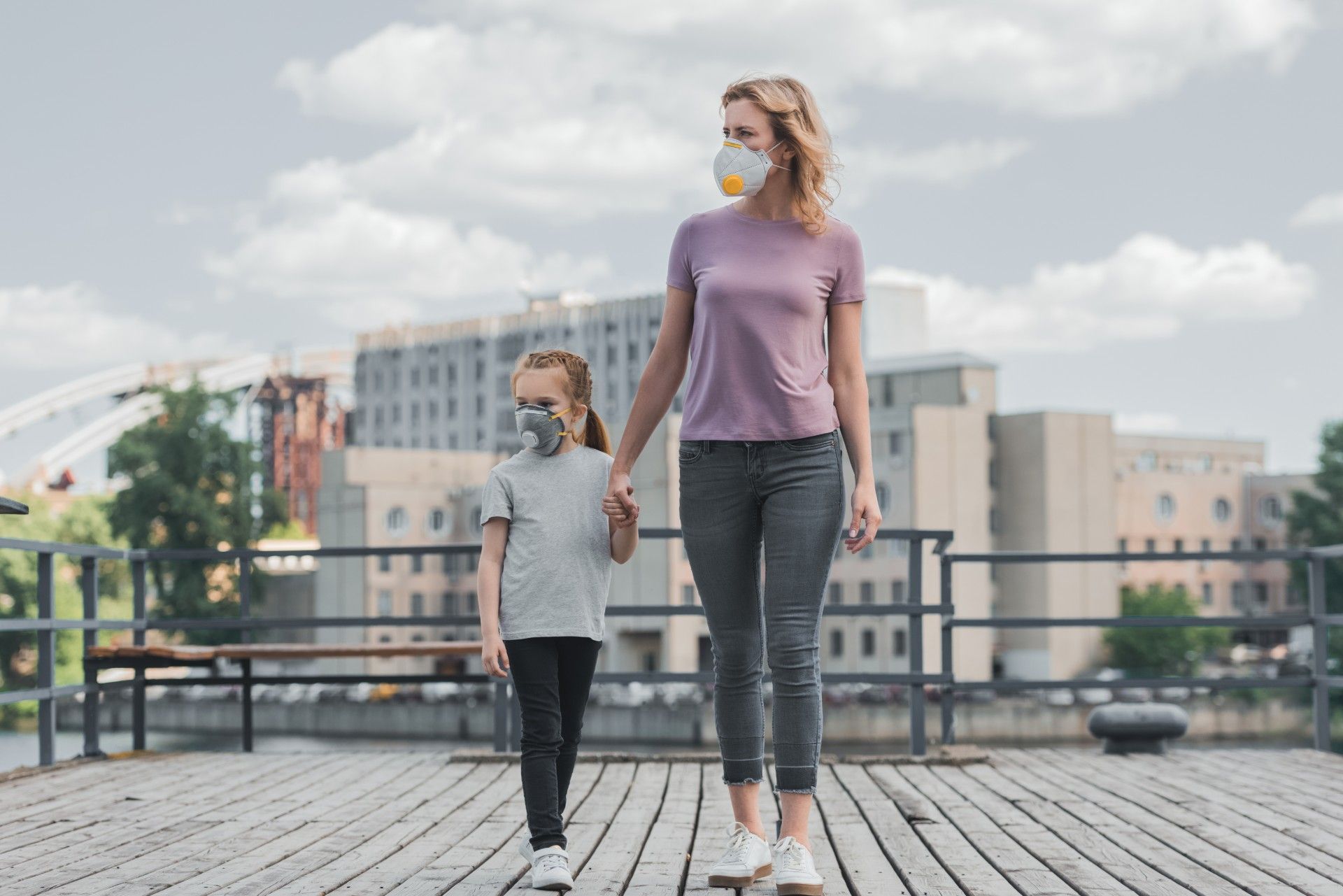 Woman and girl wear face masks and walk holding hands while it's cloudy outside - COVID-19 deaths