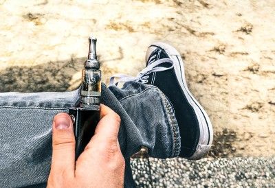 Closeup of a hand holding a vape pen near a leg wearing grey jeans and black Converse - vape samples