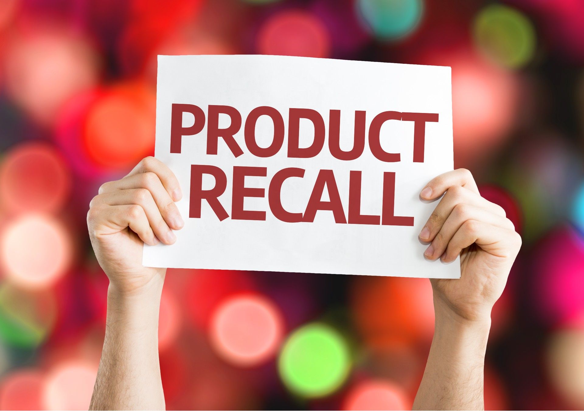 Closeup of a person's hands holding up a sign that reads "Product recall"; blurry colored lights in the background - baker ross toys