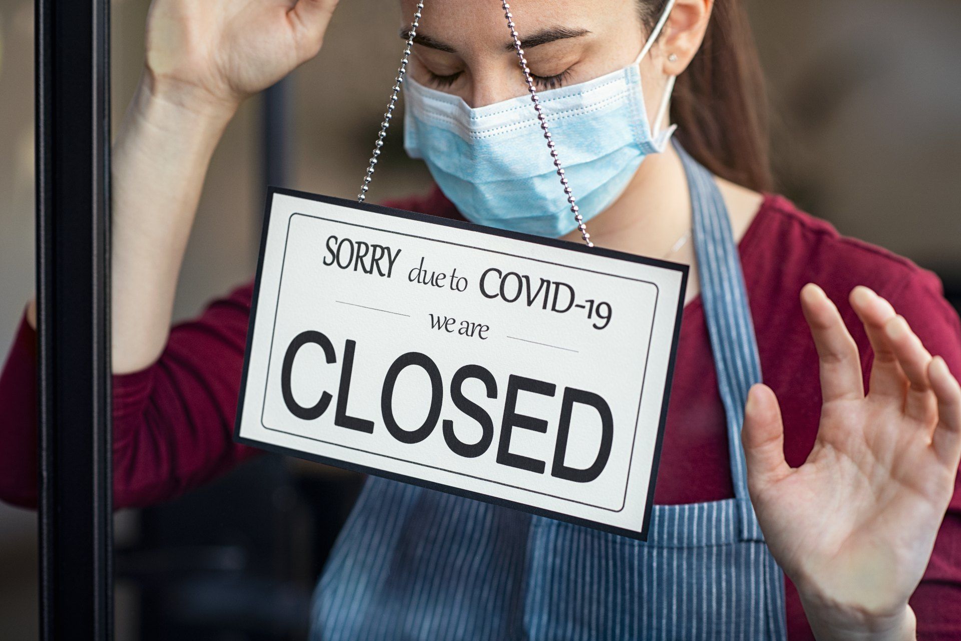 Woman in mask inside restaurant stands behind window sign that reads "sorry, due to covid-19 we are closed" - three-tier restrictions