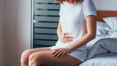 A red-haired woman sits on the edge of her bed in a white t-shirt, grabbing her abdomen in pain - royal derby hospital
