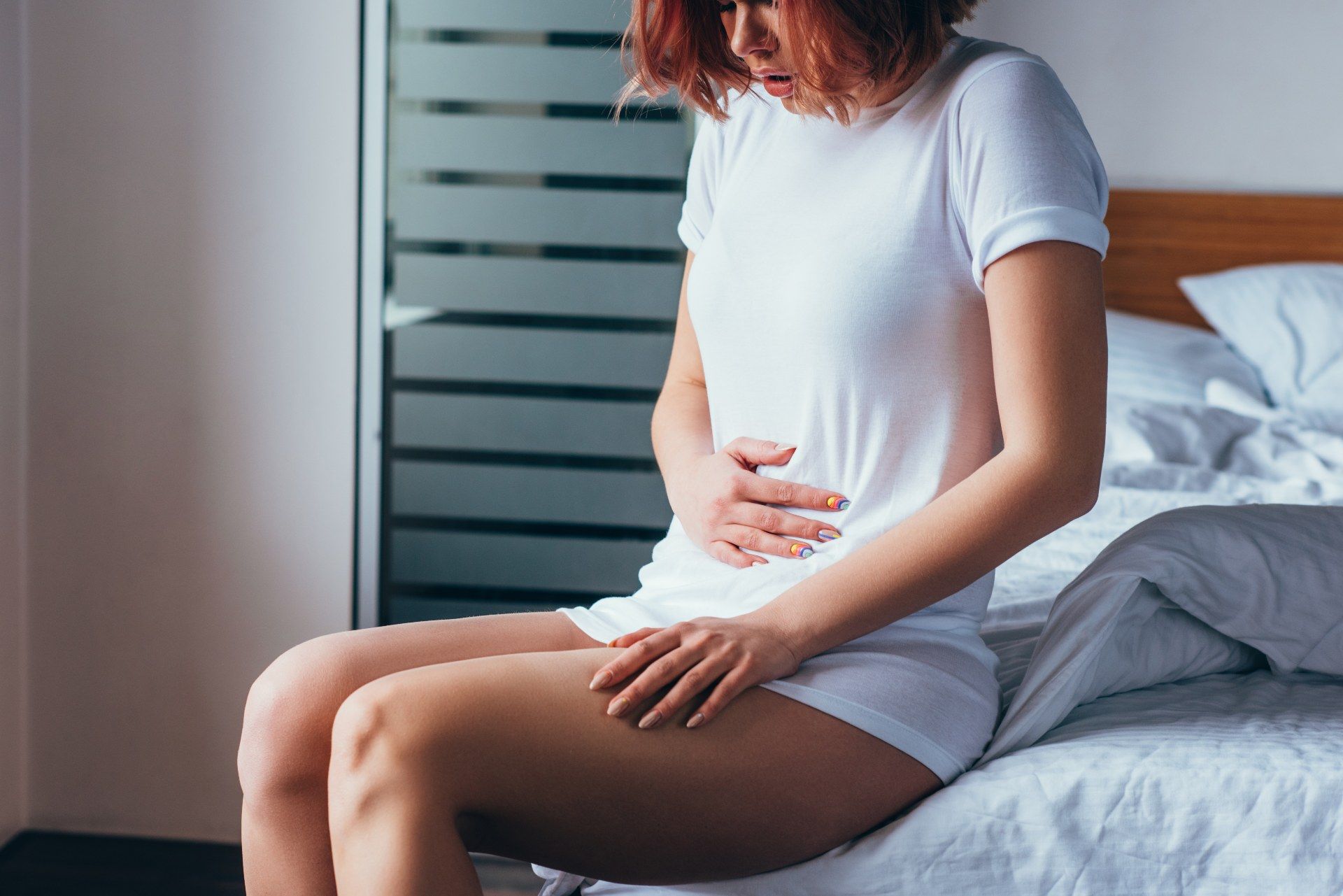 A red-haired woman sits on the edge of her bed in a white t-shirt, grabbing her abdomen in pain - royal derby hospital