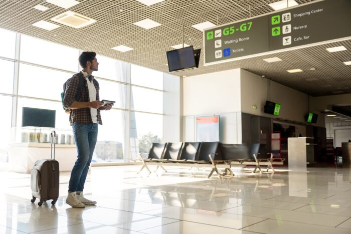 Serious young man is looking for right direction on information board at the airport. He is standing near his suitcase