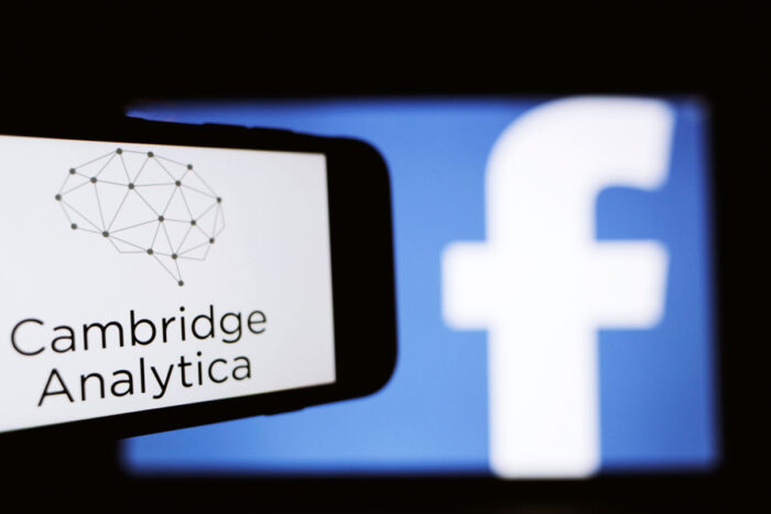 The logo of the strategic communication company Cambridge Analytica is seen on the screen of an iPhone in front of a computer screen showing Facebook logo.